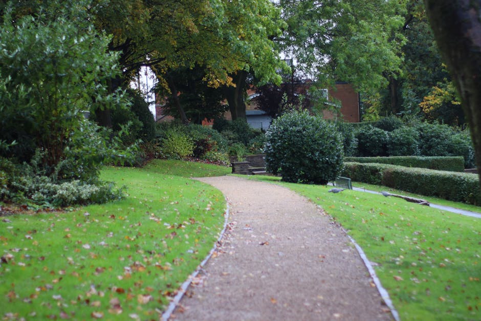 A detailed view of a well-maintained garden from an elevated perspective, showcasing lush green grass borders, neatly trimmed hedges, and vibrant flower beds along a curved stone pathway. The garden f